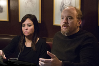 A man with a goatee gestures while talking to a woman sitting beside him in a dimly lit room.