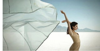 A woman in a tan dress poses with arms raised in a white salt flat under a clear sky.