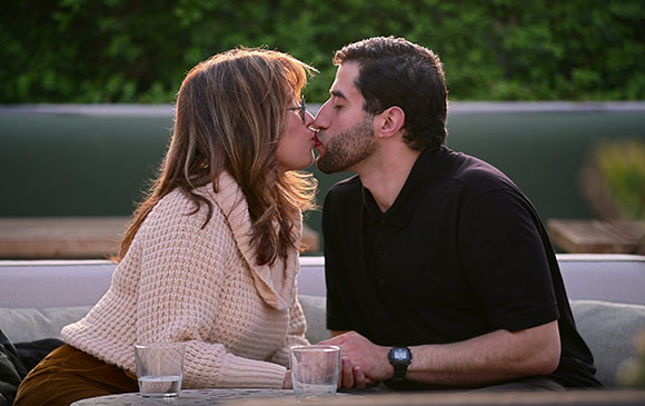 A man in a black polo and a woman in a cream sweater kiss while sitting on an outdoor sofa.