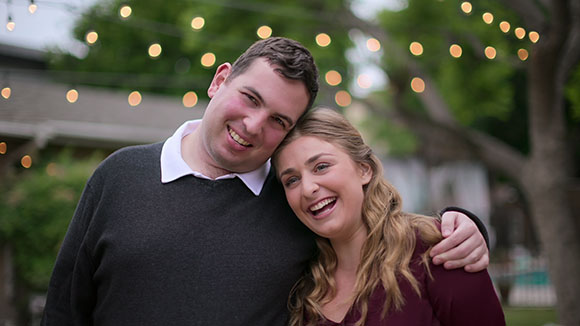A smiling man in a dark sweater and a woman in a maroon top lean their heads together.