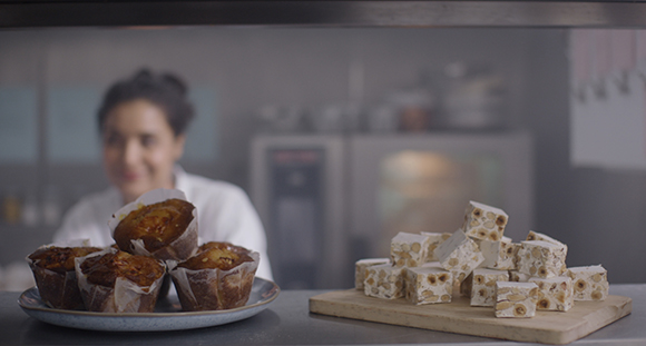 Muffins in paper liners and cubes of white nougat with nuts sit on a counter in front of a blurred person.