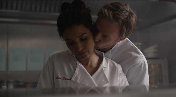 A man in a white chef coat leans toward a woman in a white chef coat in a dimly lit kitchen.