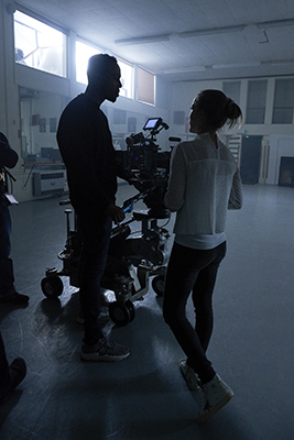 A man and a woman stand in silhouette next to a large motion picture camera on a wheeled dolly in a studio.