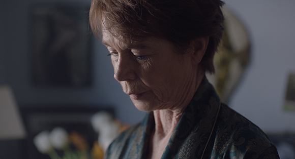 A woman with short brown hair looks downward with a serious, contemplative expression in dim lighting.