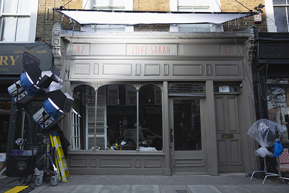 A gray storefront with 'LOVE SARAH' and '82' written on it, surrounded by film lighting equipment.