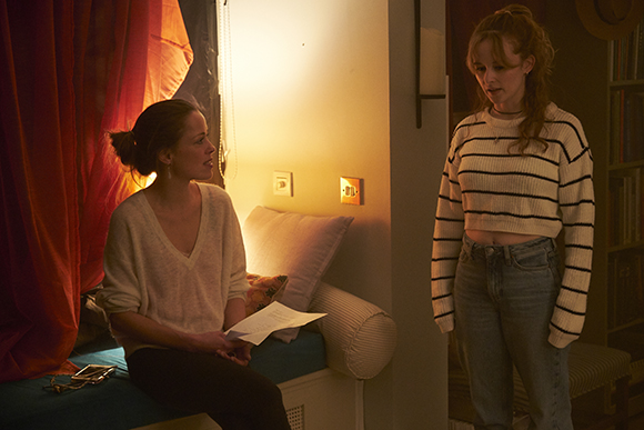 A woman sits on a bench holding paper while another woman in a striped sweater stands nearby.