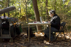 A man wearing headphones sits at a small table using a laptop in a wooded area next to a production cart.