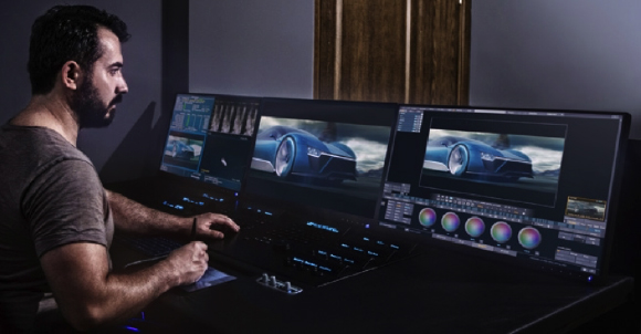 A man sits at a desk with three large monitors displaying video editing software and car footage.