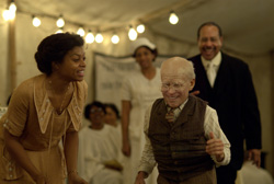 An elderly man in a vest dances while a woman in a gold dress and a man in a suit watch him smile.