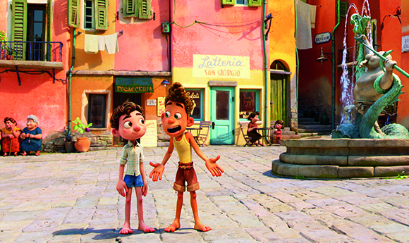 Two animated boys stand in a sunlit cobblestone plaza surrounded by colorful buildings and a fountain.