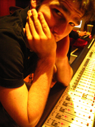 A man rests his chin on his hands while leaning over a large audio mixing console in a dimly lit room.