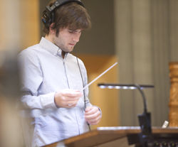 A man in a light blue shirt wearing headphones looks down at a paper near a desk lamp.