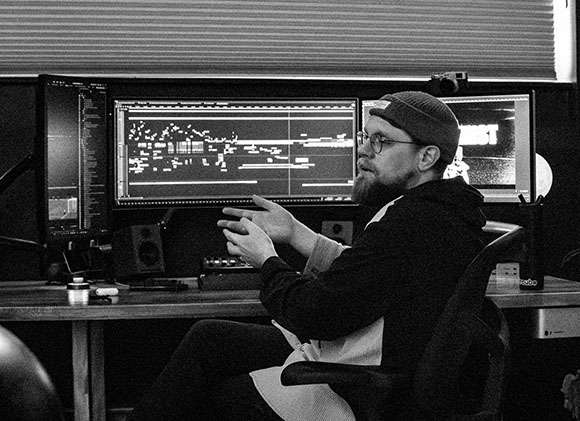 A bearded man in a beanie and glasses sits at a desk with two large computer monitors displaying digital waveforms.