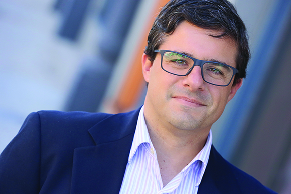 A man with dark hair and glasses wearing a navy blazer and striped shirt, smiling at the camera.