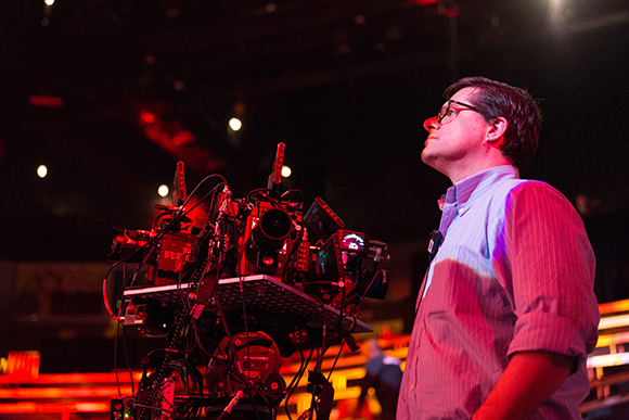 A man in glasses and a button-down shirt looks upward next to a large professional film camera rig.