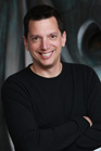 A smiling man with short dark hair wearing a black long-sleeved shirt and crossing his arms.