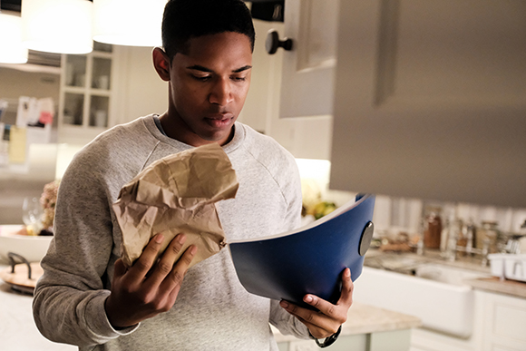 A young man in a grey sweatshirt holds a brown paper bag and a blue folder while looking down in a kitchen.