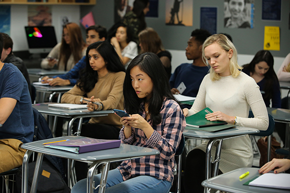 Several students sit at desks in a classroom, some looking down at books or devices.