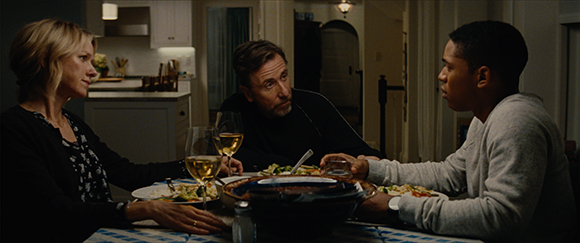 A woman, a man, and a young man sit around a dining table with food and wine in a dimly lit room.