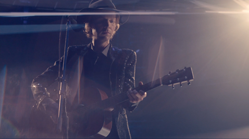 A person in a hat and sequined jacket plays an acoustic guitar on a dimly lit stage.