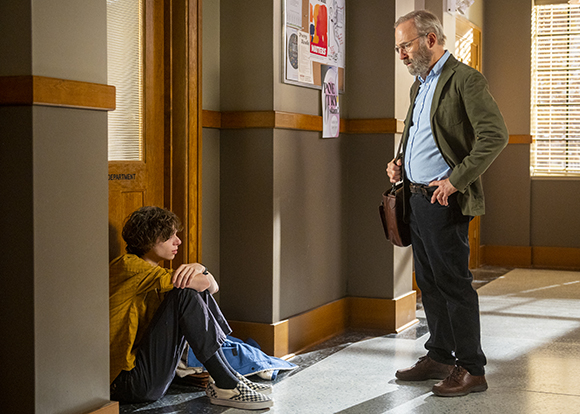 An older man in a green blazer looks down at a young man sitting on the floor in a hallway.