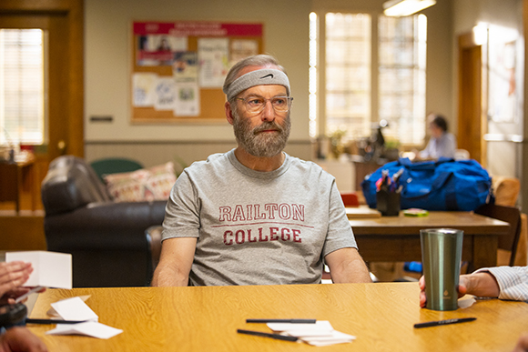 A bearded man wearing a grey Railton College t-shirt and a headband sits at a wooden table.