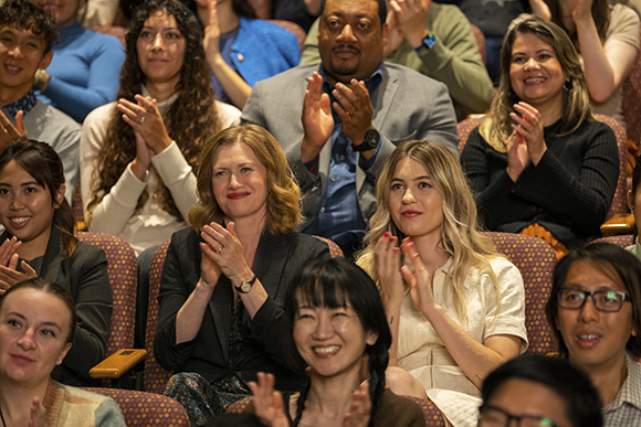 A diverse group of people sitting in theater seats, smiling and clapping their hands.