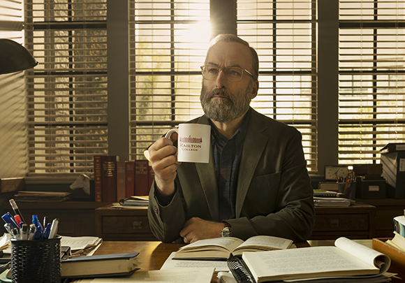 A man with glasses and a beard sits at a desk, holding a white mug in front of window blinds.