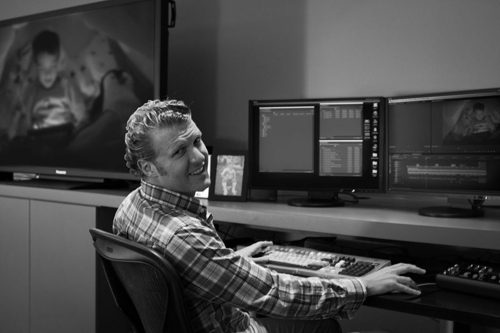 A man in a plaid shirt sits at a desk with several computer monitors in a black and white photo.