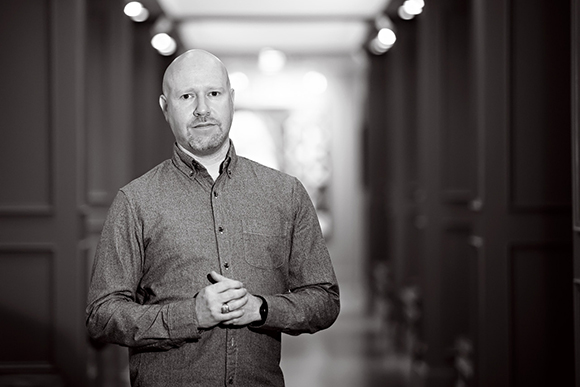A bald man in a button-down shirt stands with hands clasped in a blurred hallway.