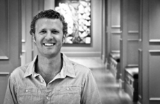 Black and white portrait of a smiling man in a collared shirt standing in a hallway.