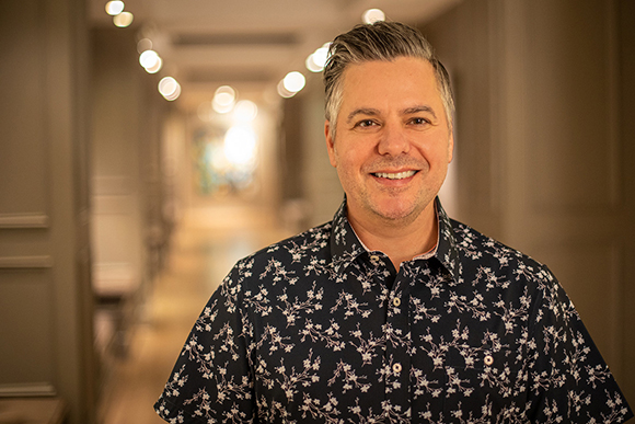 A smiling man with salt-and-pepper hair wearing a dark navy floral button-down shirt.
