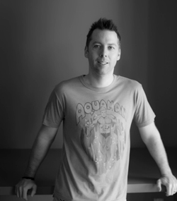 A man in a short-sleeved Aquaman graphic t-shirt stands leaning against a table.