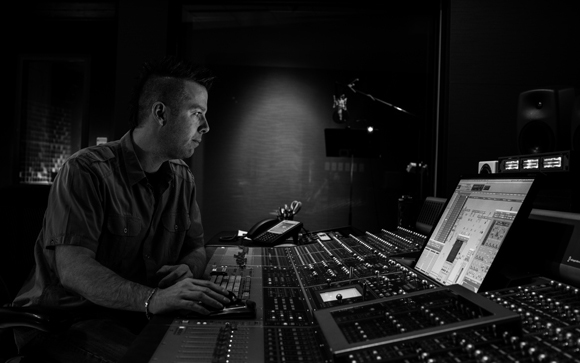 A man sits at a large professional audio mixing console in a dark recording studio.