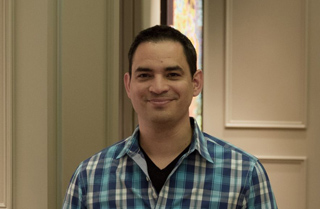 A man with short dark hair wearing a blue and white plaid shirt, smiling at the camera.