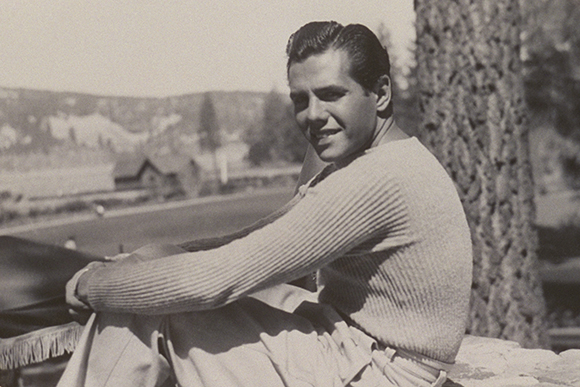 A man in a long-sleeved ribbed sweater sits outdoors with a tree and hills in the background.