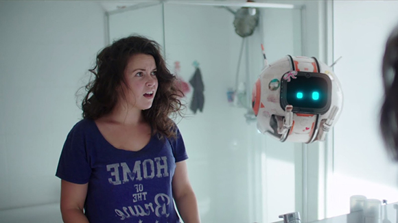A woman with curly hair looks surprised at a small, white, spherical floating robot with glowing blue eyes.