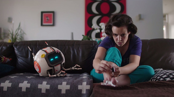 A woman in a purple shirt and teal pants sits on a leather couch next to a small, round white robot.