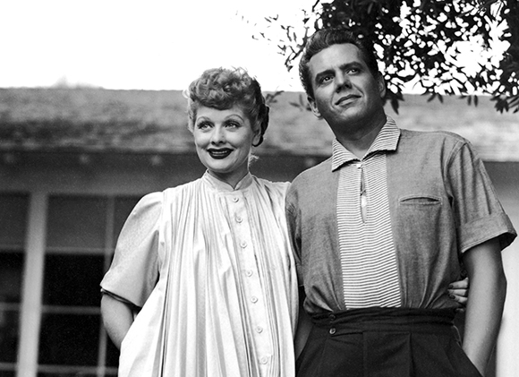 A smiling woman in a pleated dress stands next to a man in a striped polo shirt outdoors.