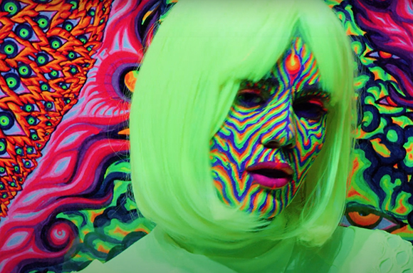 Person with neon green hair and colorful wavy face paint against a psychedelic background.