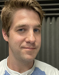 A man with light brown hair and blue eyes looking toward the camera against a dark, textured background.