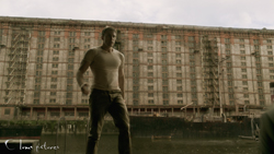 A man in a white shirt and dark pants stands in front of a massive, multi-story apartment building.