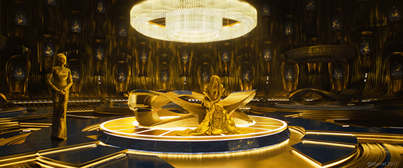 A woman stands to the left of a central platform where another figure sits in a grand, golden circular room.