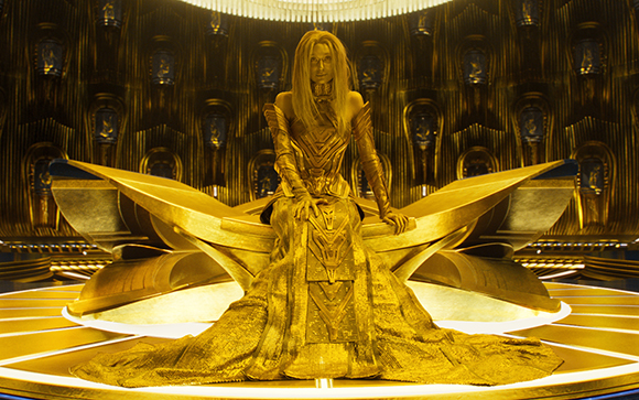 A woman with long blonde hair wears an ornate golden gown and sits on a large, sculptural golden throne.