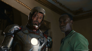 A man in silver and red metallic armor looks forward beside a man in a green shirt.