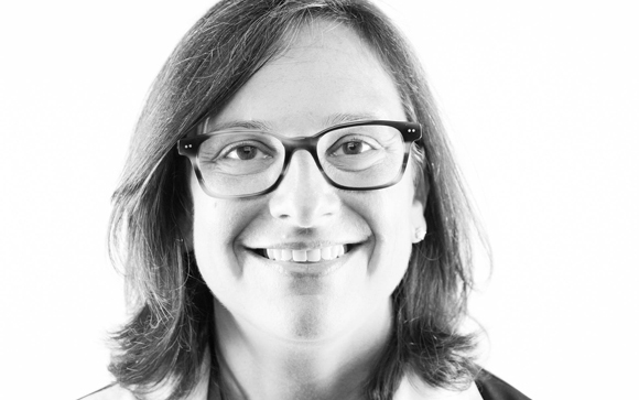 Black and white close-up portrait of a smiling woman with shoulder-length hair and dark-rimmed glasses.