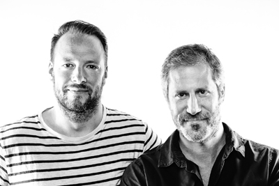 Black and white portrait of two men with facial hair standing side by side against a white background.