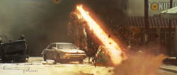 A giant creature breathes a stream of fire down a city street past parked cars.