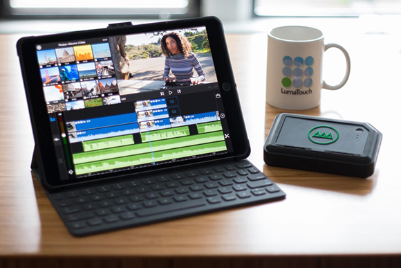 A tablet displaying video editing software next to a white mug and a small black electronic device on a wooden surface.