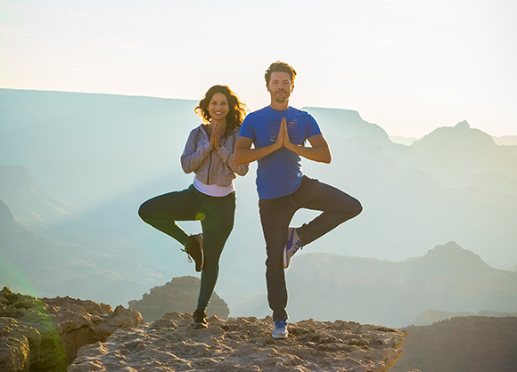 A man and woman in athletic wear perform tree pose yoga on a rocky cliff overlooking a canyon at sunrise.